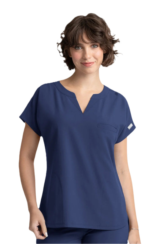 A woman wearing a navy blue Maevn Momentum Dolman 3 Pocket Scrub Top with a notched neckline, dolman sleeves, and side pockets.