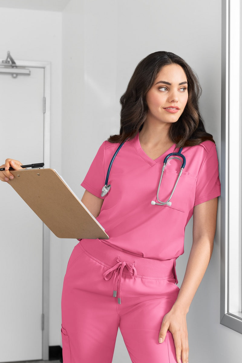 Professional healthcare worker wearing Women's Koi Cureology Aura C100 tuck in scrub top in carnation pink - moisture-wicking v-neck medical scrub with functional pockets for nurses and healthcare professionals.